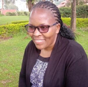 Portrait picture of Mary, the Managing Coordinator of the African Conference. Against a green background she smiles past the camera on the side. Mary wears a black and white patterned skirt and a black jacket. She wears her long black hair in many braids and wears brown glasses.