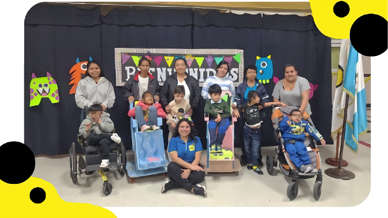 A group photograph of adults and young children in specialized adaptive chairs and strollers gathered in front of a "Bienvenidos" banner decorated with cartoon monsters. A woman in a blue shirt sits cross-legged on the floor in front of the group.